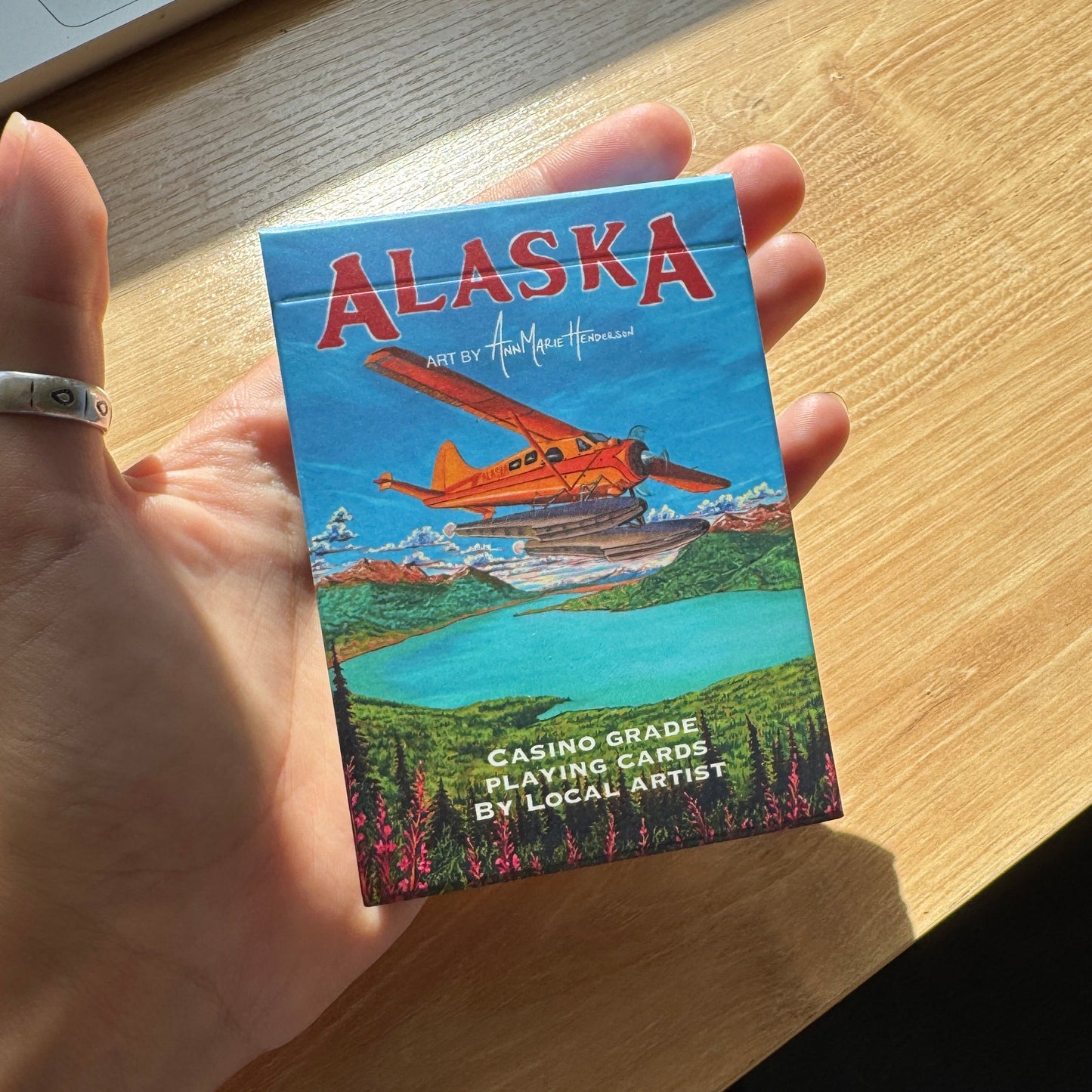 A hand holding a small printed card or book cover titled "ALASKA" with a colorful illustration of a red seaplane over a lake and forest, photographed on a wooden table in natural light.