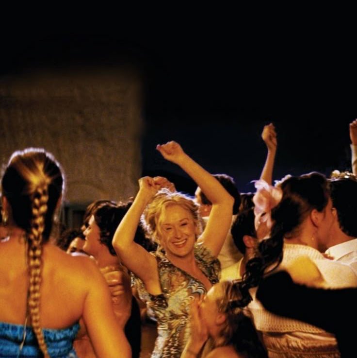 A nighttime crowd scene with several people dancing and raising their hands under warm yellow lighting. The central figure is smiling, wearing a patterned dress, while others in the crowd wear light-colored formal or partywear.